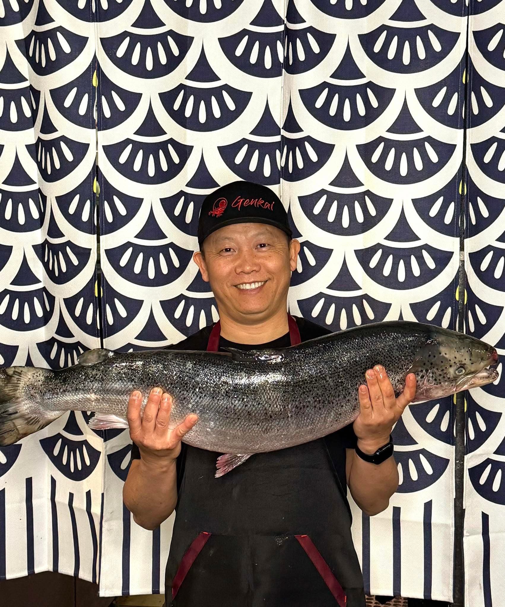 Kou Her, owner of Genkai, in a Genkai cap and apron holding a whole salmon in front of a navy-and-white noren curtain