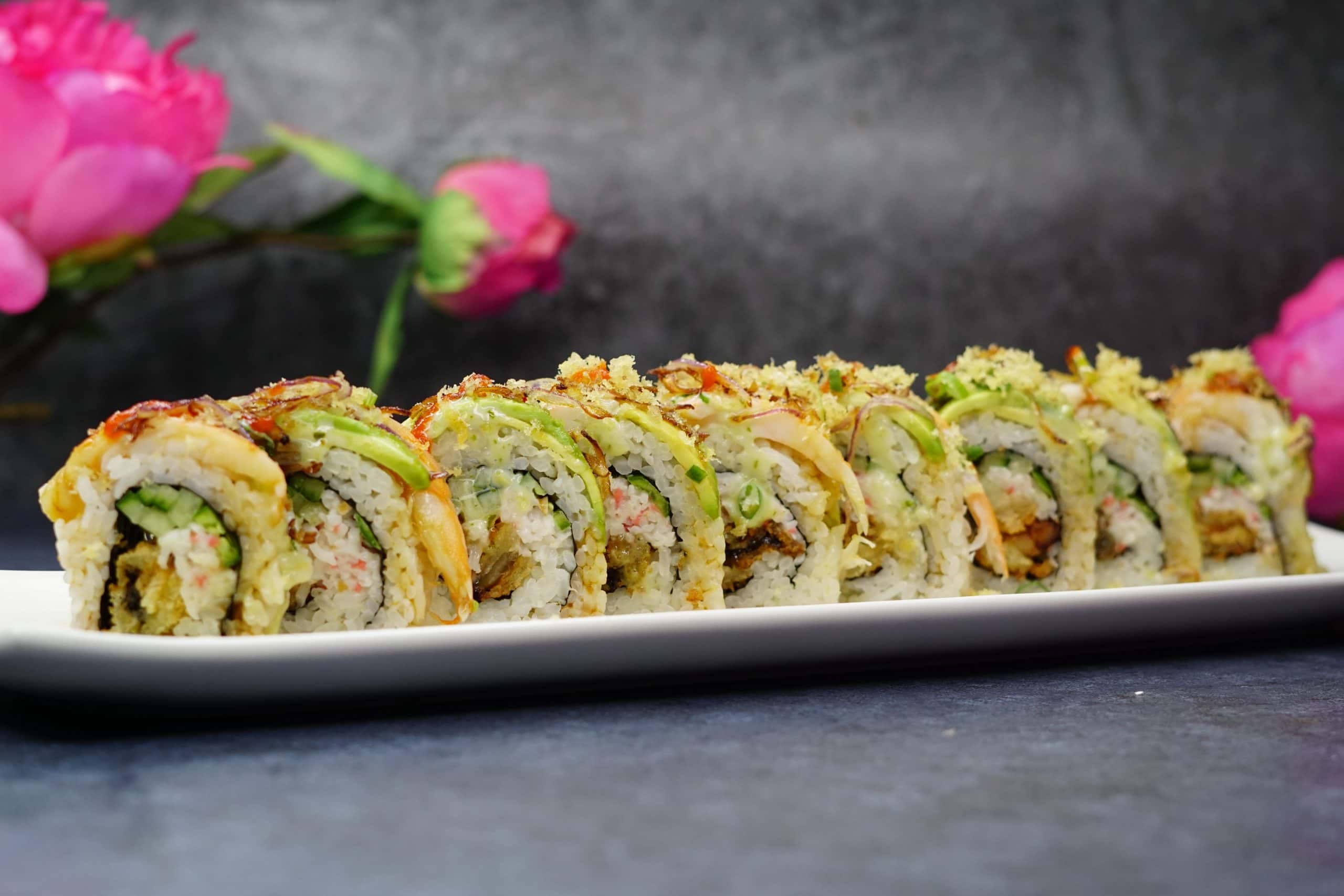 A specialty roll sliced into eight pieces, layered with avocado, crisp panko and shaved onion, with pink blossoms behind