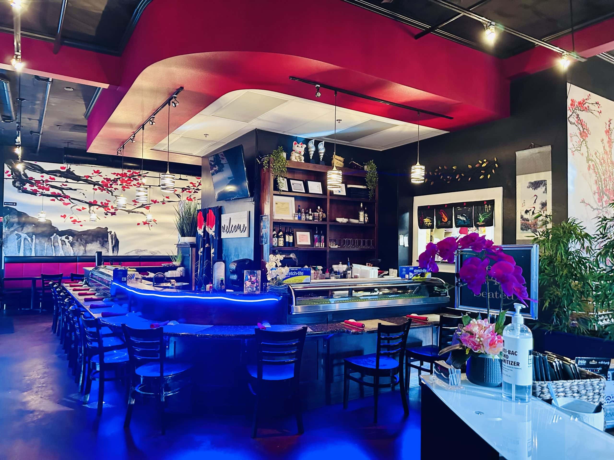 Genkai’s sushi bar lit in blue, with the refrigerated fish case, cherry blossom murals along the wall and hanging scrolls